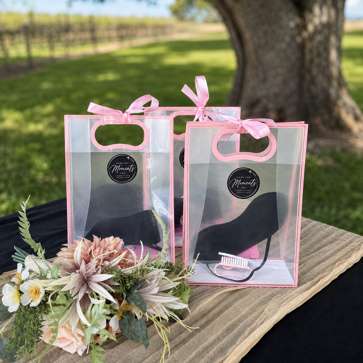 Two clear gift bags with pink trim and pink ribbons, each containing a black face mask and a pink hair tie, displayed on a table outdoors with flowers in the foreground.