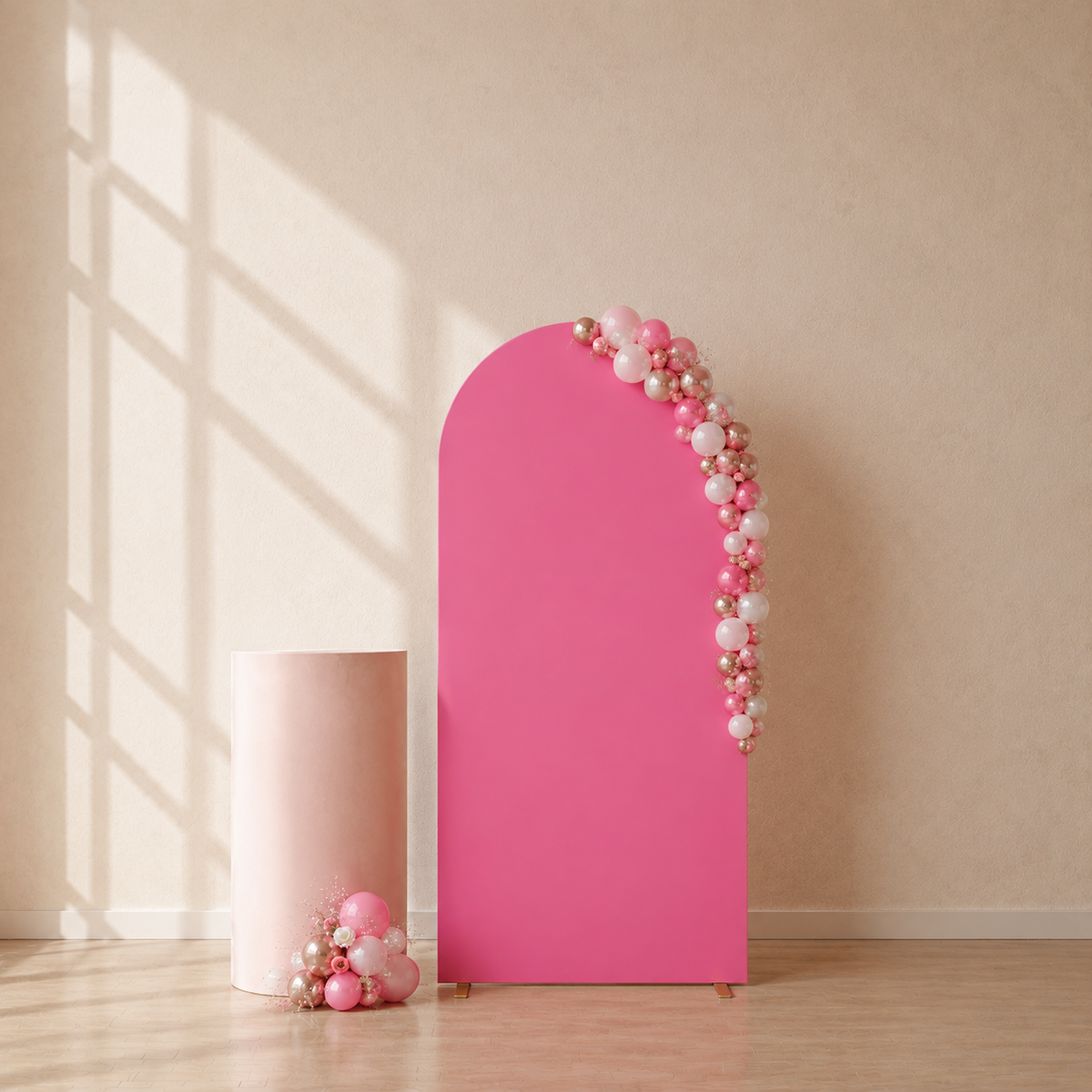 A bright pink arched backdrop stand with a balloon garland decoration in pink, white, and gold, displayed next to a matching pink cylindrical pedestal on a light wood floor against a beige wall with window shadow patterns.