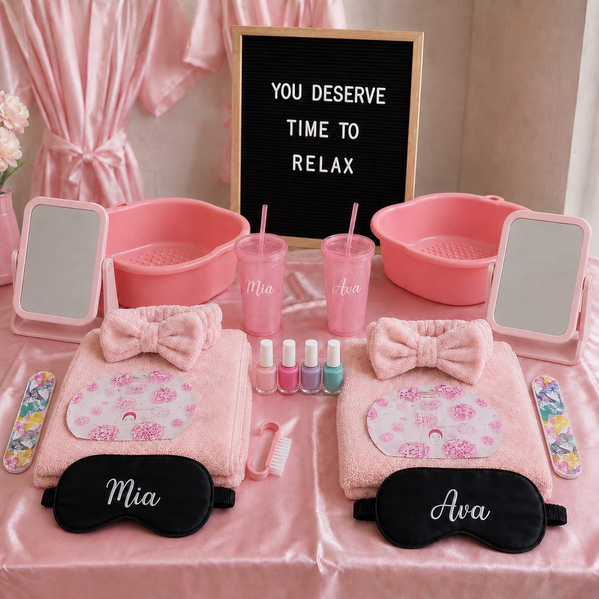 A pink spa gift set arranged on a satin-covered table, including two folded pink towels with floral eye patches, pink bow accessories, two pink tumblers with straws, two small nail polish bottles, two handheld mirrors, two pink washbasins, and two black sleep masks personalized with names. A black letter board reads 'YOU DESERVE TIME TO RELAX'.