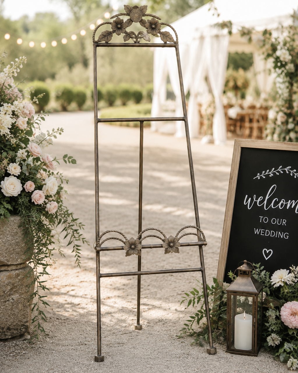 An ornate vintage-style metal wedding guest book stand in a rustic outdoor venue setting, flanked by floral arrangements and a welcome sign.