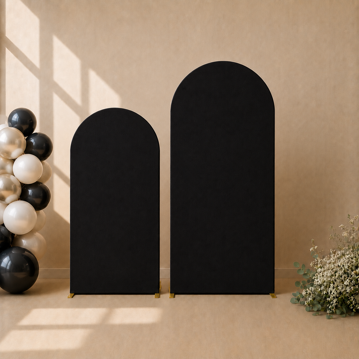 Two black arched freestanding panels or screens on a beige floor against a beige wall, styled with black, white, and metallic balloons on the left and dried flowers on the right.