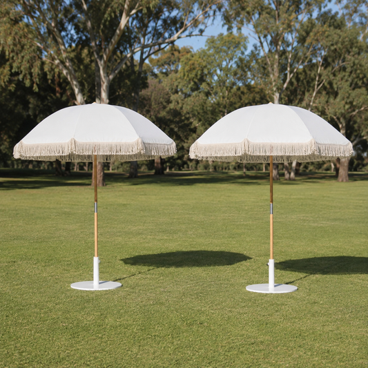 Two white patio umbrellas with fringe trim and light wood poles, standing on a green lawn with trees in the background.