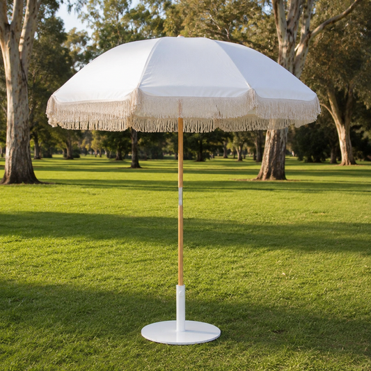 A white beach umbrella with a beige fringe trim and a light wood pole, standing on a white round base in a grassy park with trees in the background.