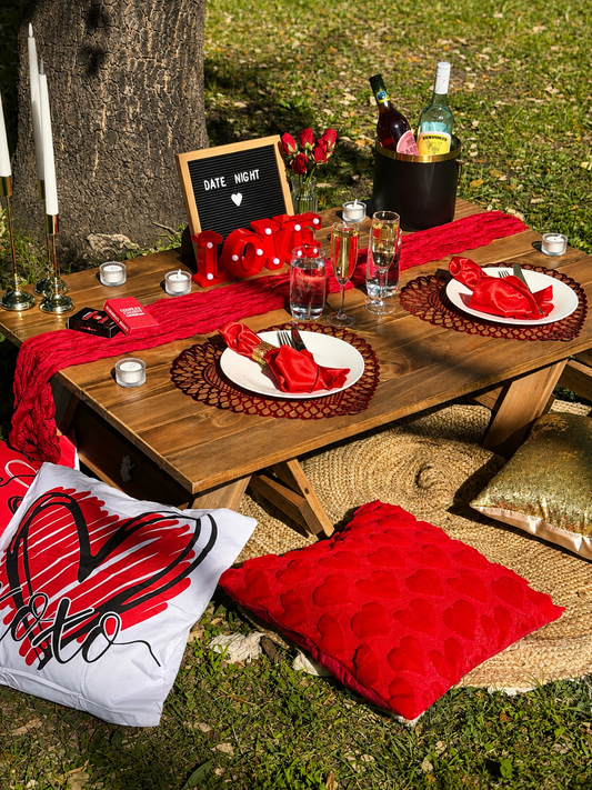 Outdoor romantic picnic setup on a wooden table with red scarves, white plates, red napkins, candles, a letter board reading 'DATE NIGHT', and red pillows on grass.