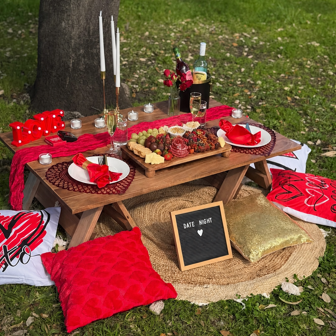 Outdoor picnic setup on a grassy lawn under a tree, featuring a wooden table with plates, candles, a charcuterie board, red napkins and tablecloths, decorative pillows, and a small chalkboard sign reading 'DATE NIGHT'.