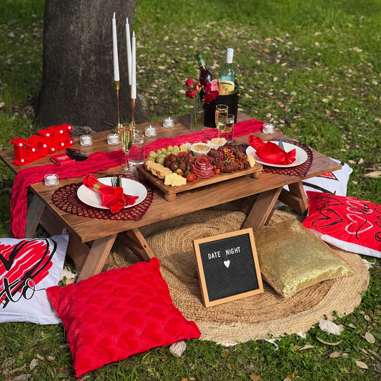 Outdoor picnic setup on a grassy lawn under a tree, featuring a wooden table with plates, candles, a charcuterie board, red napkins and tablecloths, decorative pillows, and a small chalkboard sign reading 'DATE NIGHT'.