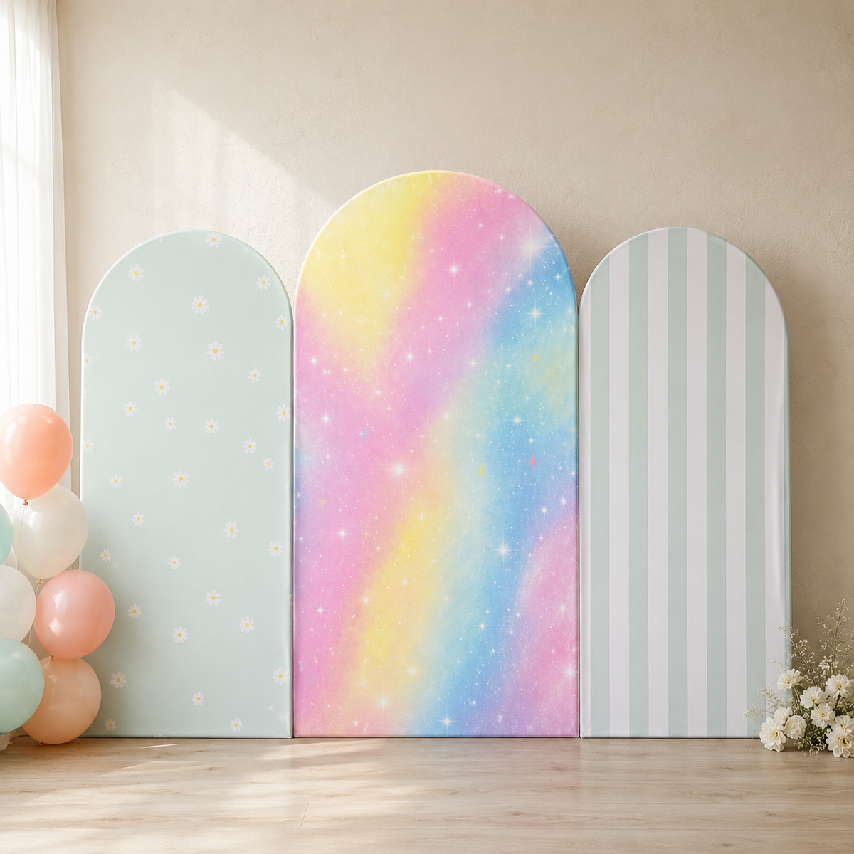 Three pastel arch-shaped party backdrop panels leaning against a beige wall on a light wood floor. The center panel has a rainbow galaxy print with pink, yellow, blue, and purple tones, while the left panel is pale mint with small white flowers, and the right panel has pale mint and white vertical stripes.
