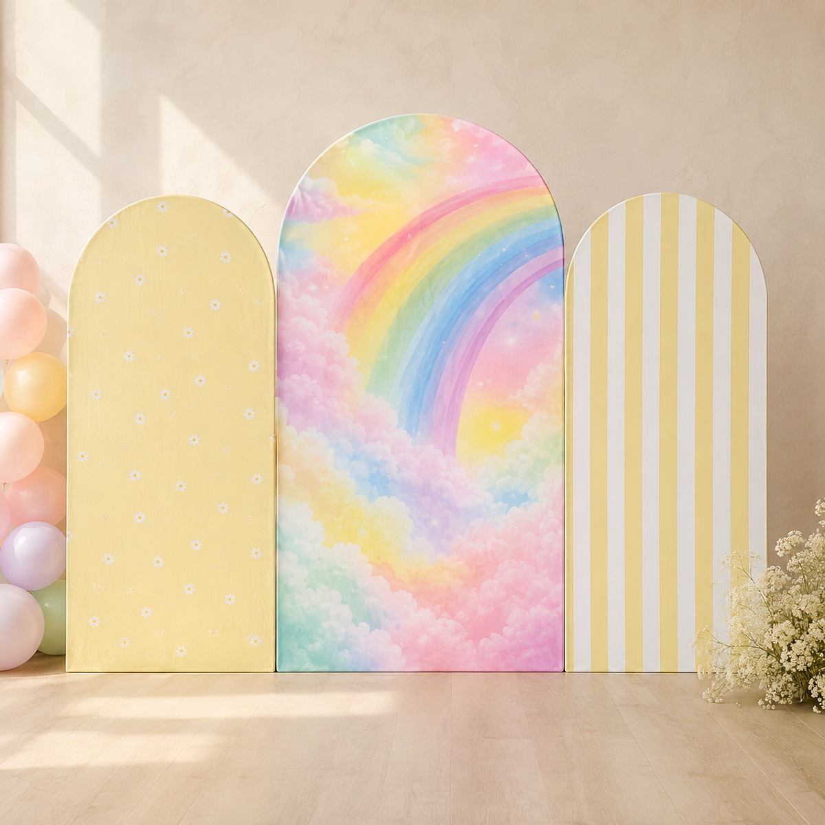 Three pastel party backdrop arches displayed on a light wood floor against a beige wall. The center arch features a colorful pastel rainbow and clouds design, flanked by a pale yellow arch with white dots on the left and a pale yellow-and-white striped arch on the right.