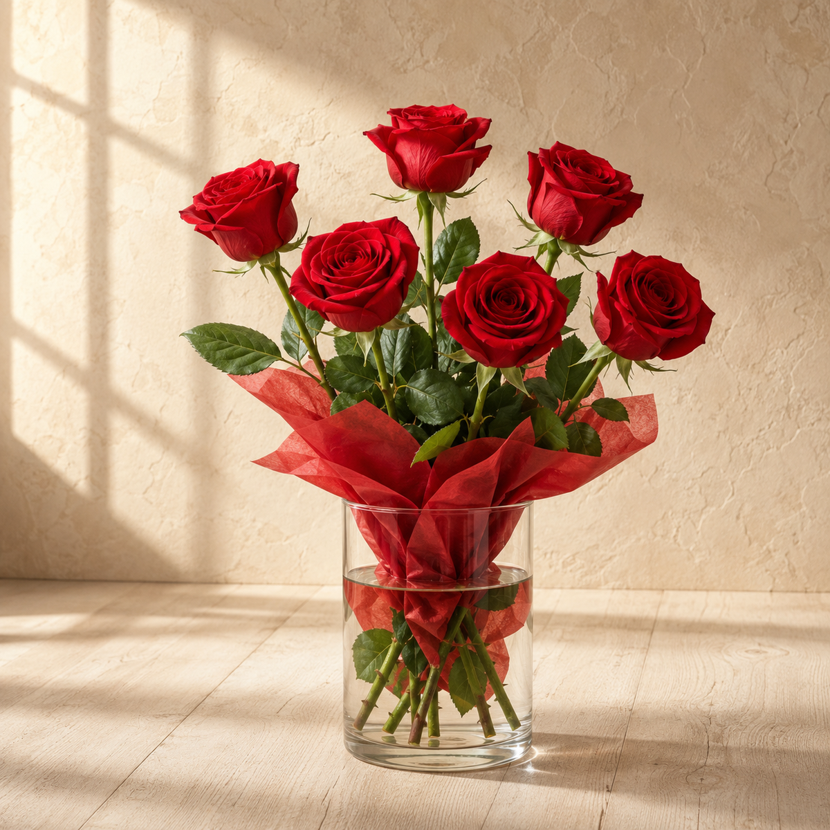 A bouquet of seven red roses with green leaves and stems, wrapped in red tissue paper and arranged in a clear glass vase on a light wood floor against a textured beige wall with soft window shadows.