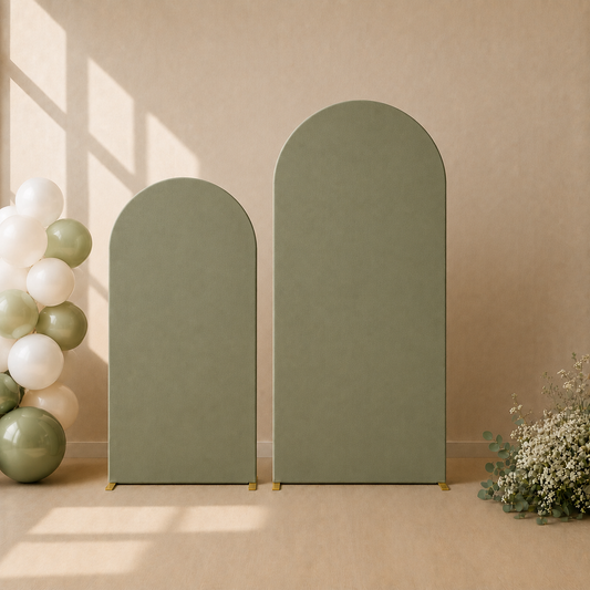 Two tall arched foam party backdrops in a muted sage green color, standing on a beige floor against a beige wall with decorative balloons and flowers.