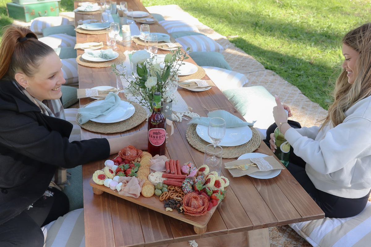 Two women sitting at opposite ends of a long wooden outdoor dining table set with plates, napkins, and a charcuterie board. The woman on the left wears a black jacket, and the woman on the right wears a white sweatshirt.