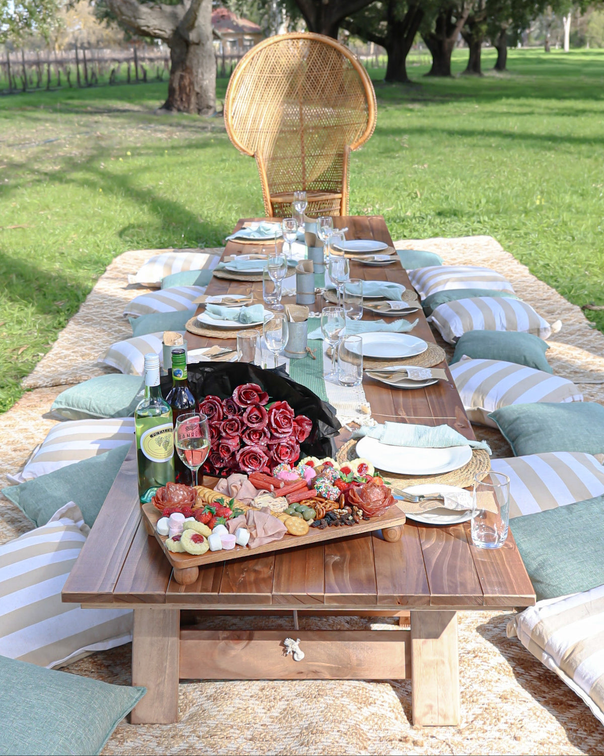 An outdoor picnic setup on a wooden table with plates, glasses, a bottle of wine, and a large basket of red roses, surrounded by striped cushions on a grassy lawn with trees in the background.