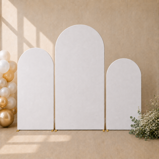 Three white arched photo backdrops of different heights arranged on a beige floor against a textured beige wall, with gold balloon accents on the left and white flowers on the right.